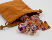 Load image into Gallery viewer, a clear set of dice with orange and purple swirled in. D20 with AbleGamers joystick logo. all the numbers are in black spiling out of a light brown velvet bag