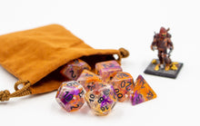 Load image into Gallery viewer, a clear set of dice with orange and purple swirled in. D20 with AbleGamers joystick logo. all the numbers are in black spiling out of a light brown velvet bag. Small D&D Mini in the background.
