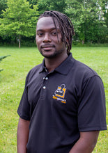 Load image into Gallery viewer, Jamile, a black-American with dreads, wearing a black polo shirt with a collar and 3 white buttons. On the left chest is the AbleGamers logo, an 80s joystick on an orange square