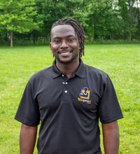 Load image into Gallery viewer, Jamile, a black-American with dreads, with a big smile, wearing a black polo shirt with a collar and 3 white buttons. On the left chest is the AbleGamers logo, an 80s joystick on an orange square