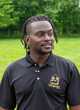 Load image into Gallery viewer, Jamile, a black-American with dreads, looking away, wearing a black polo shirt with a collar and 3 white buttons. On the left chest is the AbleGamers logo, an 80s joystick on an orange square