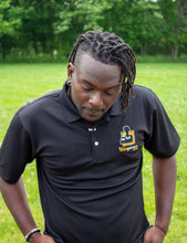 Load image into Gallery viewer, Jamile, a black-American with dreads, looking down, hands in pocket, wearing a black polo shirt with a collar and 3 white buttons. On the left chest is the AbleGamers logo, an 80s joystick on an orange square