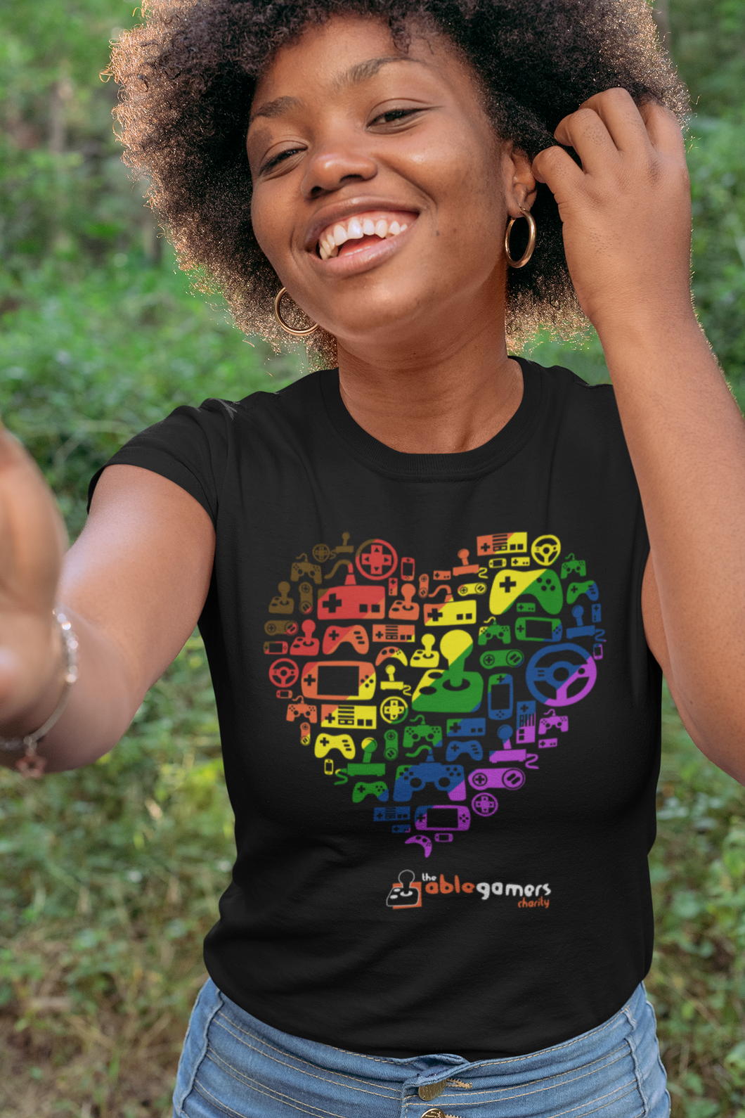 An African-American women wearing a black t-shirt. On the shirt is a heart made up of the AbleGamers logo, and came controller icons. The heart is colored in a rainbow, starting with Brown in the upper left to purple in the lower right. AbleGamers is at the bottom, and 
