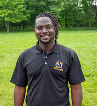 Load image into Gallery viewer, Jamile, a black-American with dreads, with a big smile, wearing a black polo shirt with a collar and 3 white buttons. On the left chest is the AbleGamers logo, an 80s joystick on an orange square
